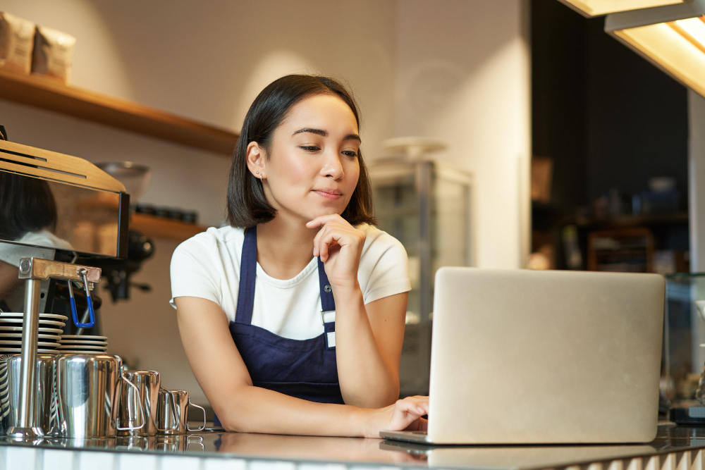 Barista works on laptop at coffee shop counter, thoughtful expression